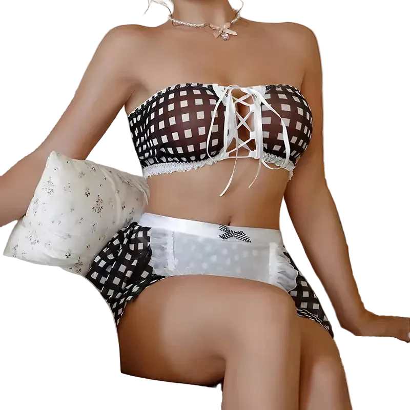 A woman seated on a striped sofa wearing a black-and-white checkered lingerie set with white lace trim and a lace-up front. A teddy bear and pillows are visible in the background.