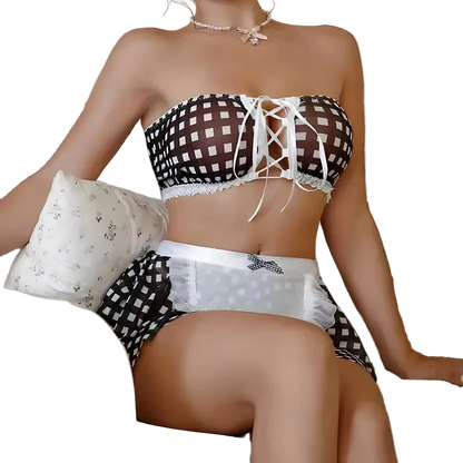 A woman seated on a striped sofa wearing a black-and-white checkered lingerie set with white lace trim and a lace-up front. A teddy bear and pillows are visible in the background.