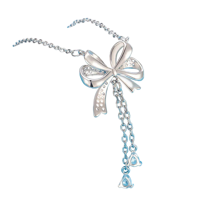 A silver-toned bow pendant necklace with dangling chain and clear teardrop-shaped stones, displayed on a light blue cylindrical stand against a soft blue background.