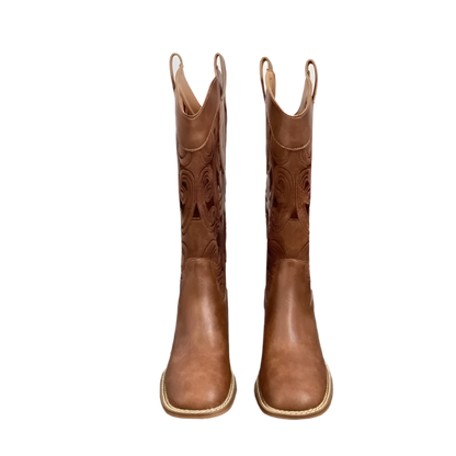 Pair of brown cowboy boots on a wooden floor with a white wall in the background.