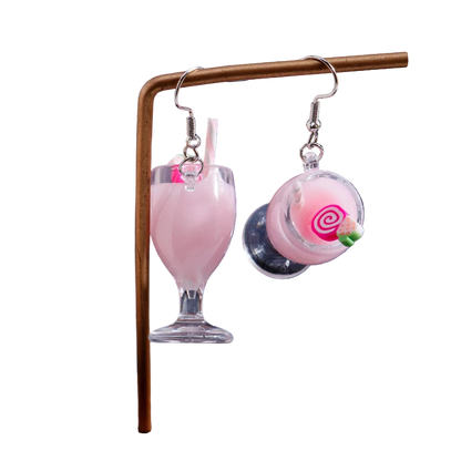 Earrings shaped like pink milkshakes with a straw on a stand against a dark background