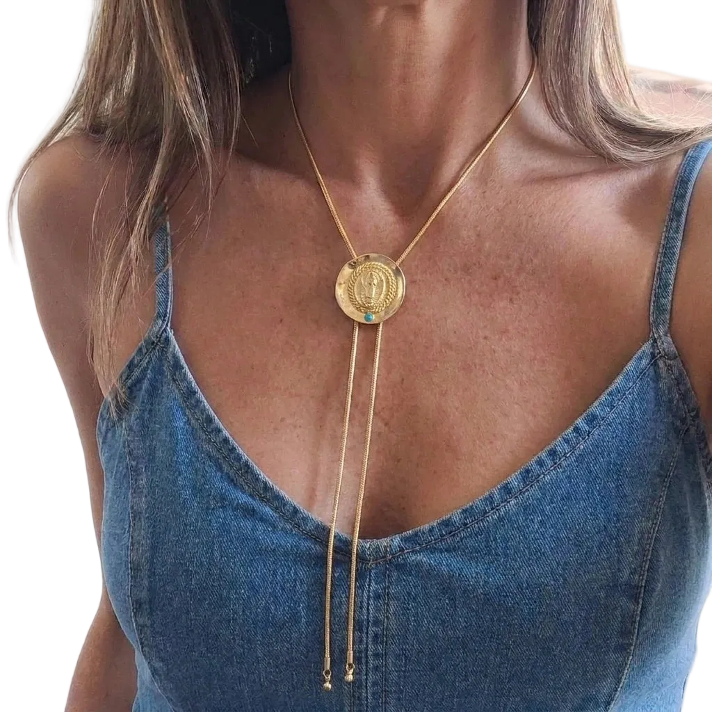 A gold-toned lariat necklace with a circular pendant featuring a religious figure, worn by a model in a blue denim camisole top.
