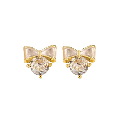 A pair of gold bow stud earrings with clear faceted round crystals, displayed on a white decorative surface.