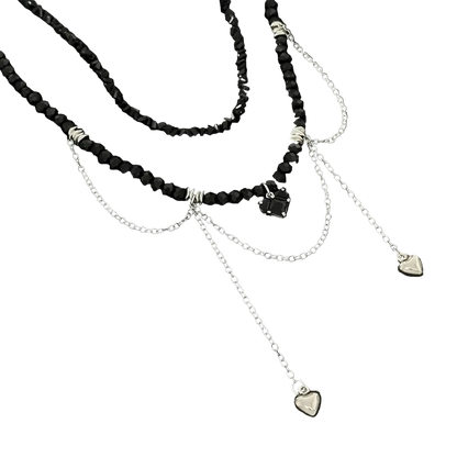 A layered necklace featuring black faceted beads, silver chain drapes, and small heart-shaped charms, displayed on a dark textured surface.