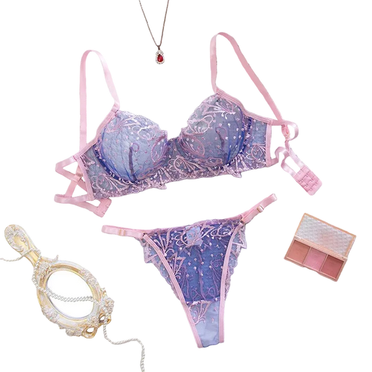 A pink and purple lace lingerie set with matching bra and panties, styled in a flat-lay arrangement on a white textured fabric background with decorative props including a small mirror, necklace, flowers, and a makeup palette.