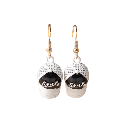 A pair of dangling earrings shaped like baseball caps, with white textured caps featuring black front panels and gold fishhook wires, displayed on a beige jewelry stand.