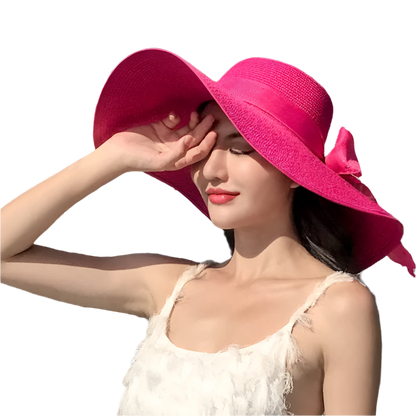 Woman wearing a bright pink hat with a large bow, standing outdoors with buildings and trees in the background.