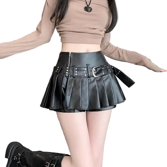 A person wearing a black faux-leather pleated mini skirt with silver buckle and stud details, paired with a beige long-sleeve crop top and black boots, standing indoors next to a suitcase.