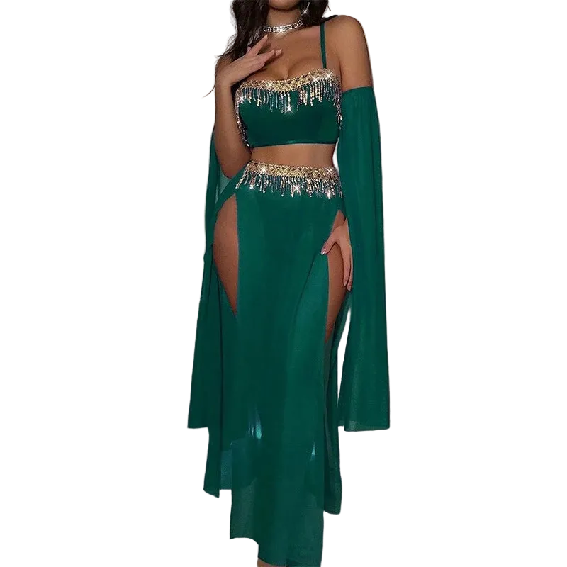A woman models a two-piece emerald green belly dance costume with gold metallic fringe details, featuring a crop top, high-slit skirt, and matching long cape sleeves, set against a dark room with a pink neon light.