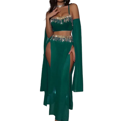A woman models a two-piece emerald green belly dance costume with gold metallic fringe details, featuring a crop top, high-slit skirt, and matching long cape sleeves, set against a dark room with a pink neon light.