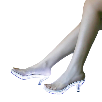 Clear high-heeled shoes with LED lights worn by a person on a reflective surface.