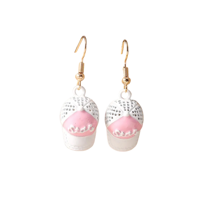 A pair of dangling earrings displayed on a beige jewelry stand. Each earring has a gold-tone fishhook wire and a white lace-like top section with a pink lower section shaped like a small cupcake or pastry.