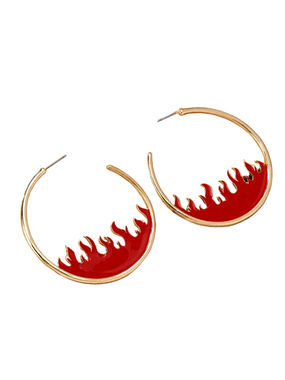 Gold hoop earrings with a red enamel lower half featuring cutout flame shapes, displayed on a wooden block over a cream satin fabric background.