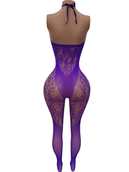 A mannequin wearing a purple lace bodysuit with matching sheer tights, shown from the back in a softly lit bedroom with a bed, lamp, and curtains in the background.