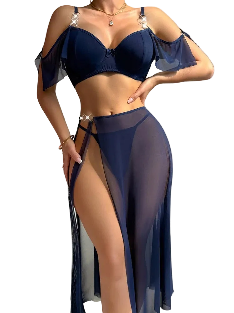 A woman modeling a dark navy blue lingerie set consisting of a bra top with sheer cold-shoulder flutter sleeves and a matching sheer high-slit skirt or drape, against a plain light gray wall.