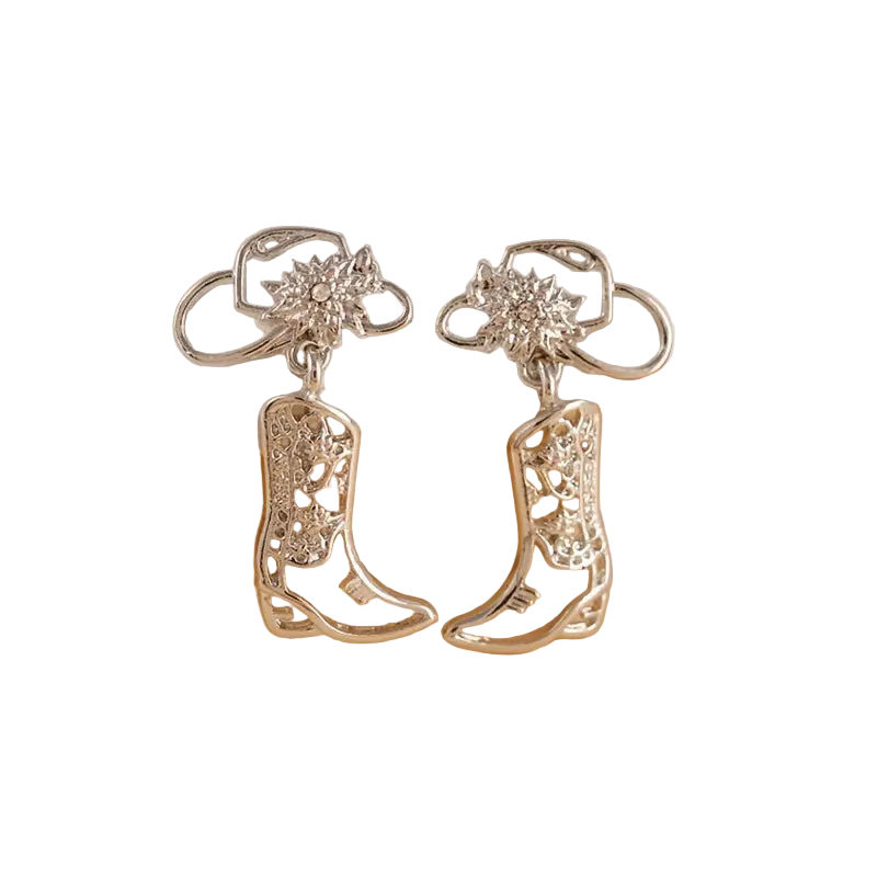 A pair of silver-toned western-style drop earrings with boot-shaped charms and floral stud tops, displayed on a curved white stand against a warm beige background.