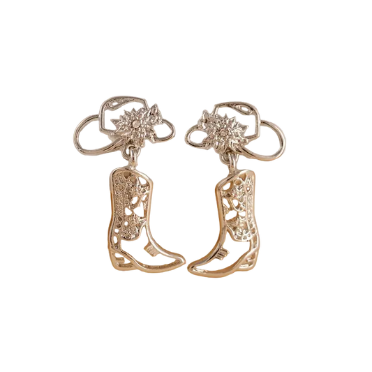 A pair of silver-toned western-style drop earrings with boot-shaped charms and floral stud tops, displayed on a curved white stand against a warm beige background.