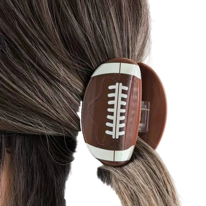 A football-shaped hair claw clip with a brown and white design, shown clipped to a person's dark hair.