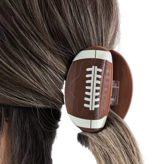 A football-shaped hair claw clip with a brown and white design, shown clipped to a person's dark hair.