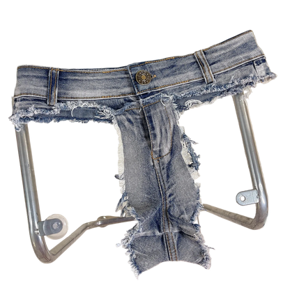 A pair of heavily distressed and ripped blue denim jeans with a frayed, raw hem and leg edges, displayed on a clear glass table with a metal frame.