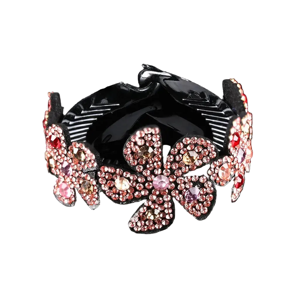 A black hair claw clip decorated with large, pink and rose gold rhinestone-embellished flower motifs, set against a solid black background with a soft reflection below.