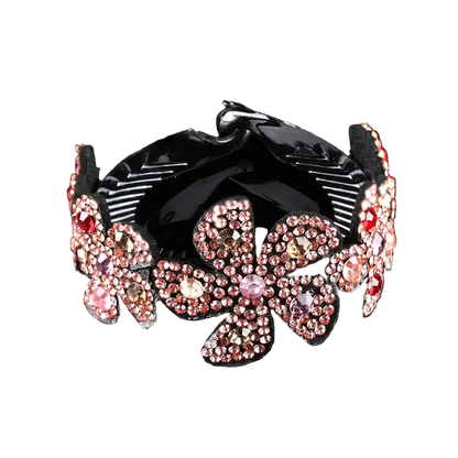 A black hair claw clip decorated with large, pink and rose gold rhinestone-embellished flower motifs, set against a solid black background with a soft reflection below.