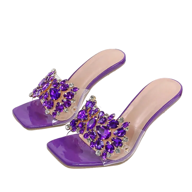 Purple mule sandals with clear acrylic block heels and a wide strap heavily embellished with purple rhinestones, displayed on a white fabric background next to a denim prop.