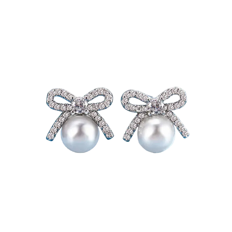 A pair of bow-shaped stud earrings with white pearl-like beads and sparkling silver-toned crystal-encrusted bows, displayed on a blue surface with a marble block in the background.