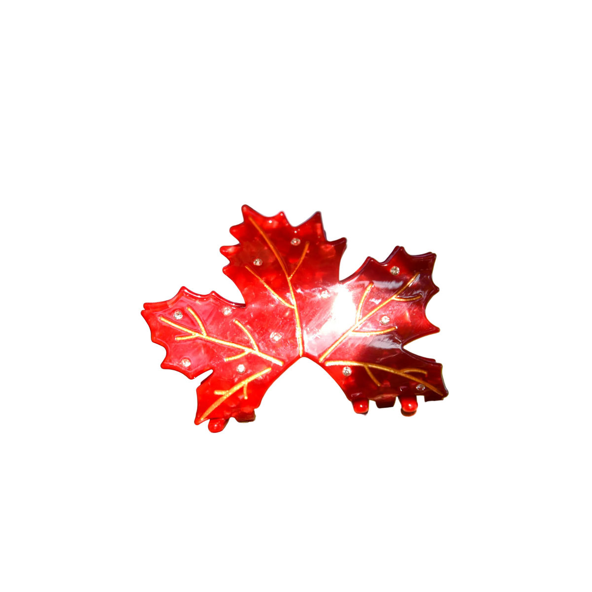A glossy red maple leaf-shaped ornament with gold vein details and small clear rhinestones, resting on a tree branch outdoors with a blurred natural background.