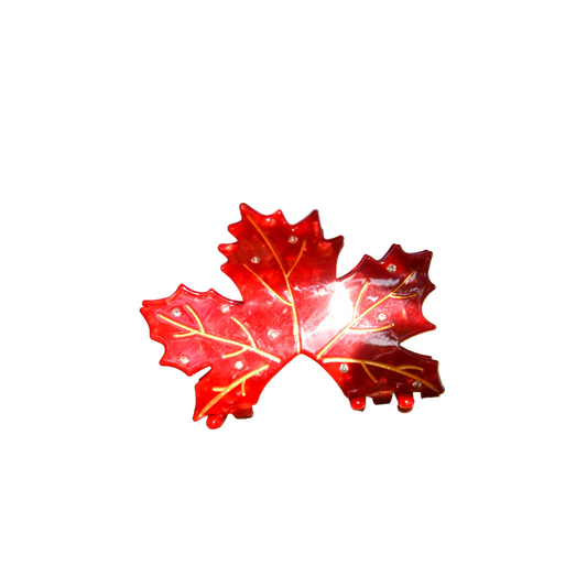 A glossy red maple leaf-shaped ornament with gold vein details and small clear rhinestones, resting on a tree branch outdoors with a blurred natural background.