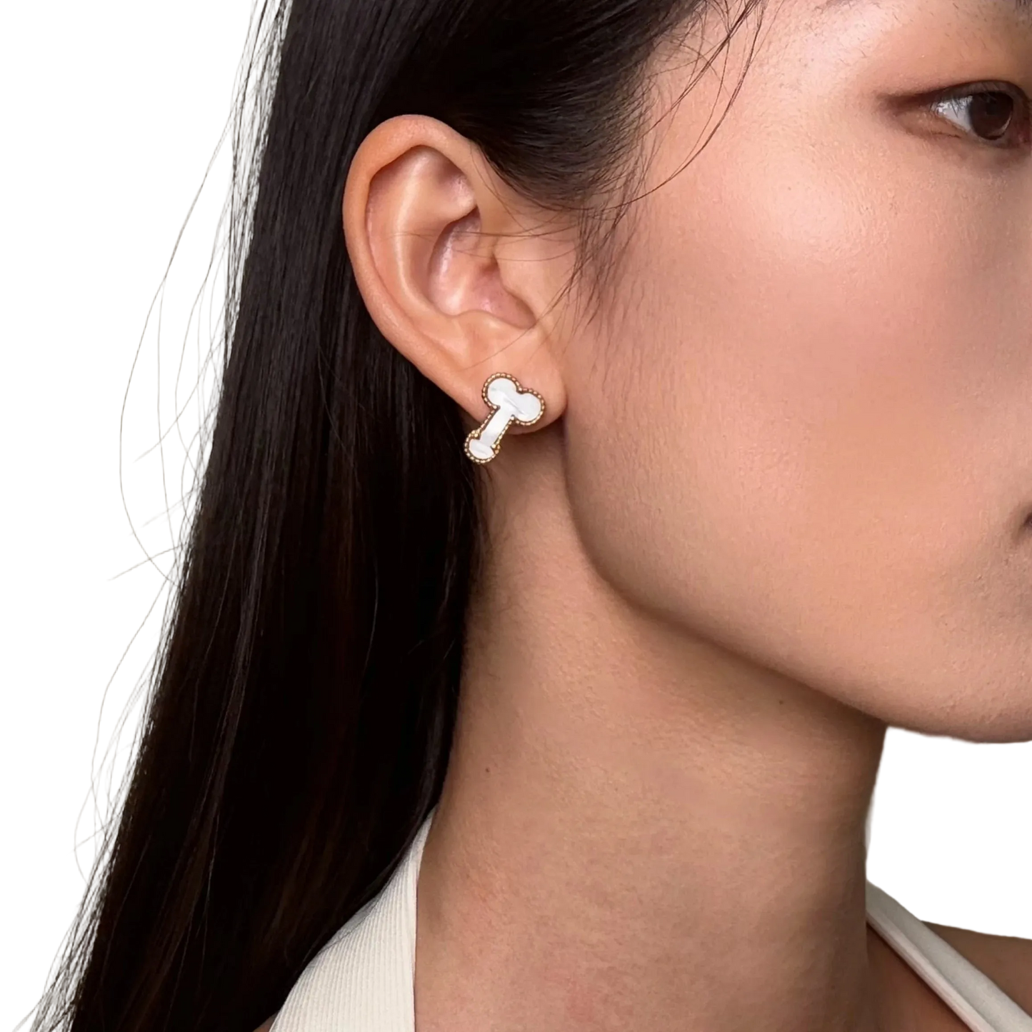 A close-up of a woman's ear wearing a small bone-shaped stud earring with a white center and silver outline, against a plain light beige background.