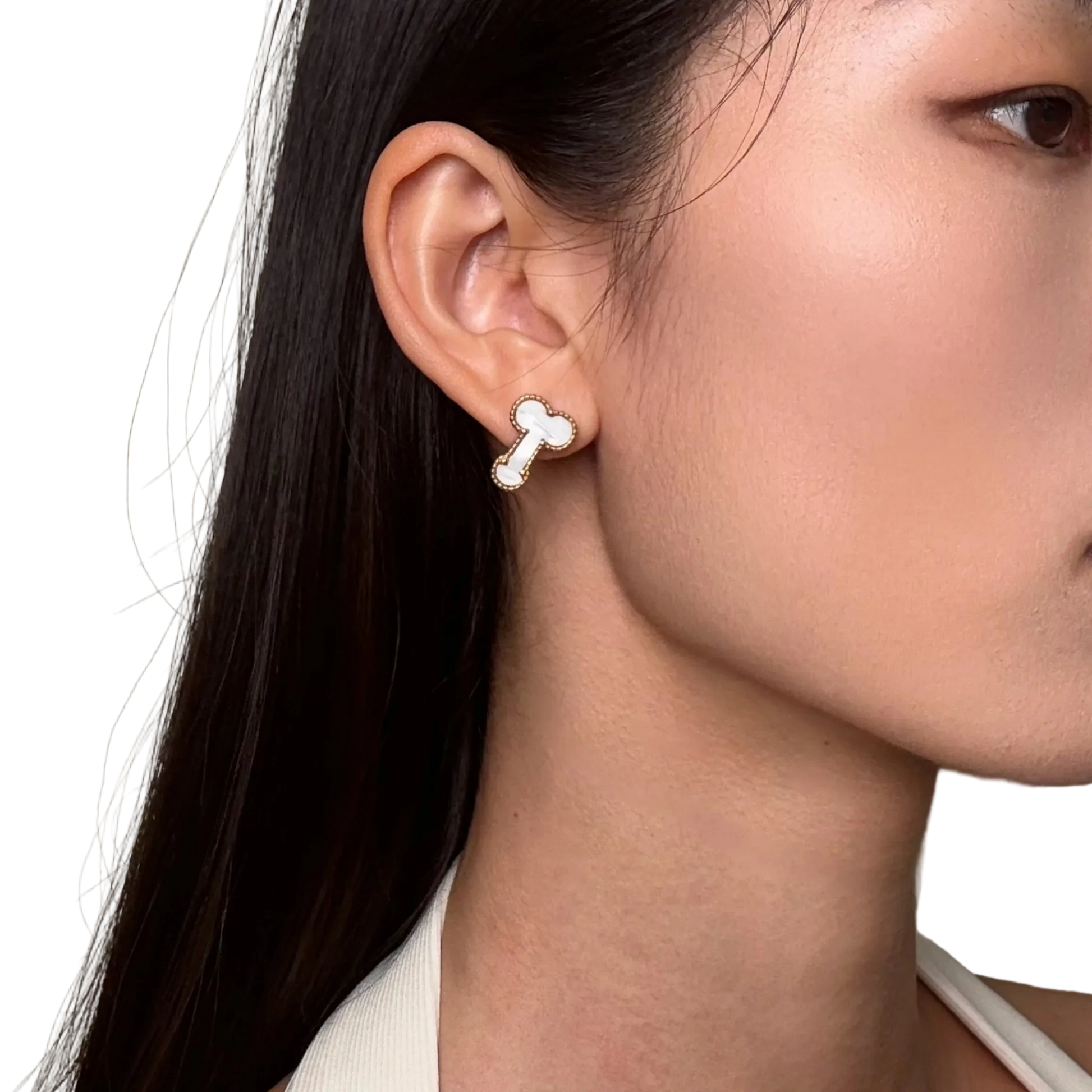 A close-up of a woman's ear wearing a small bone-shaped stud earring with a white center and silver outline, against a plain light beige background.