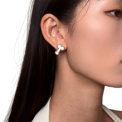 A close-up of a woman's ear wearing a small bone-shaped stud earring with a white center and silver outline, against a plain light beige background.