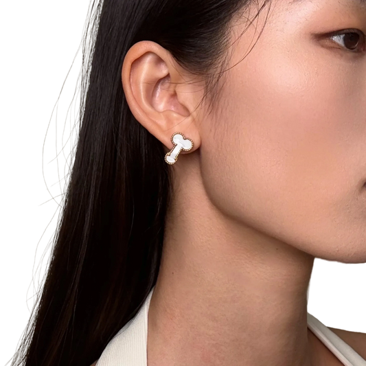 A close-up of a woman's ear wearing a small bone-shaped stud earring with a white center and silver outline, against a plain light beige background.