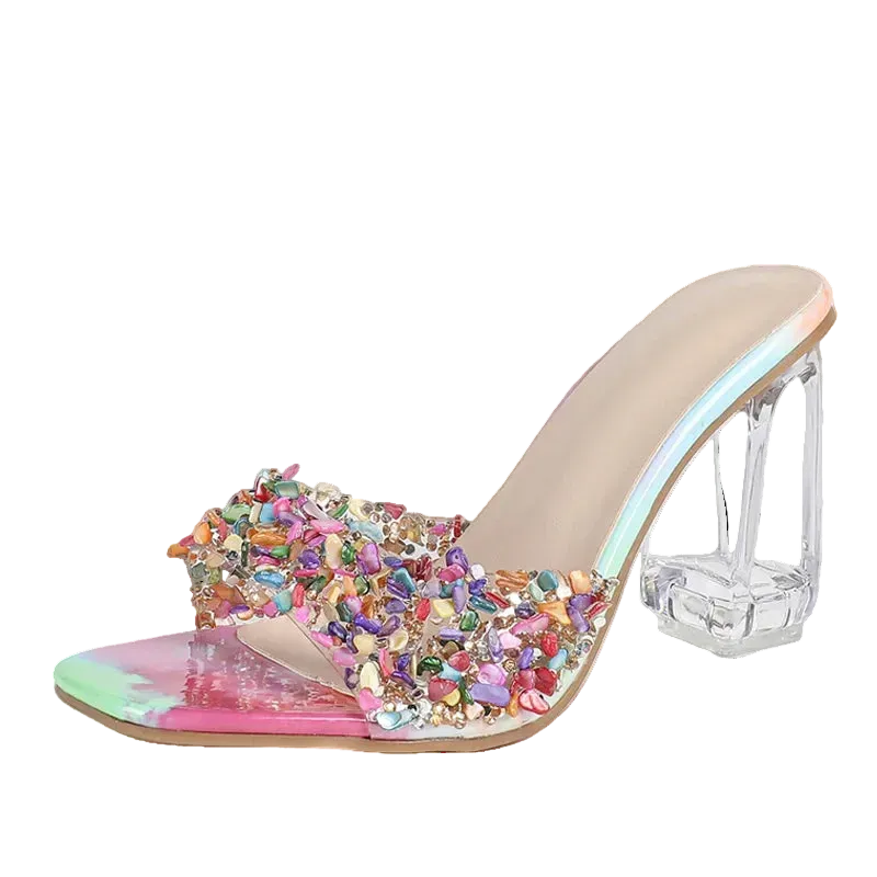 a clear acrylic block heel, a transparent strap embellished with multicolored rhinestones, and a glossy iridescent pink and green toe cap, shown on a light gray background.