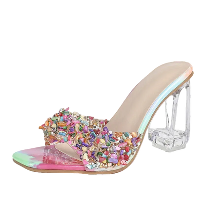 a clear acrylic block heel, a transparent strap embellished with multicolored rhinestones, and a glossy iridescent pink and green toe cap, shown on a light gray background.