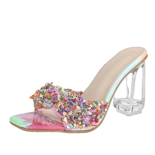 a clear acrylic block heel, a transparent strap embellished with multicolored rhinestones, and a glossy iridescent pink and green toe cap, shown on a light gray background.