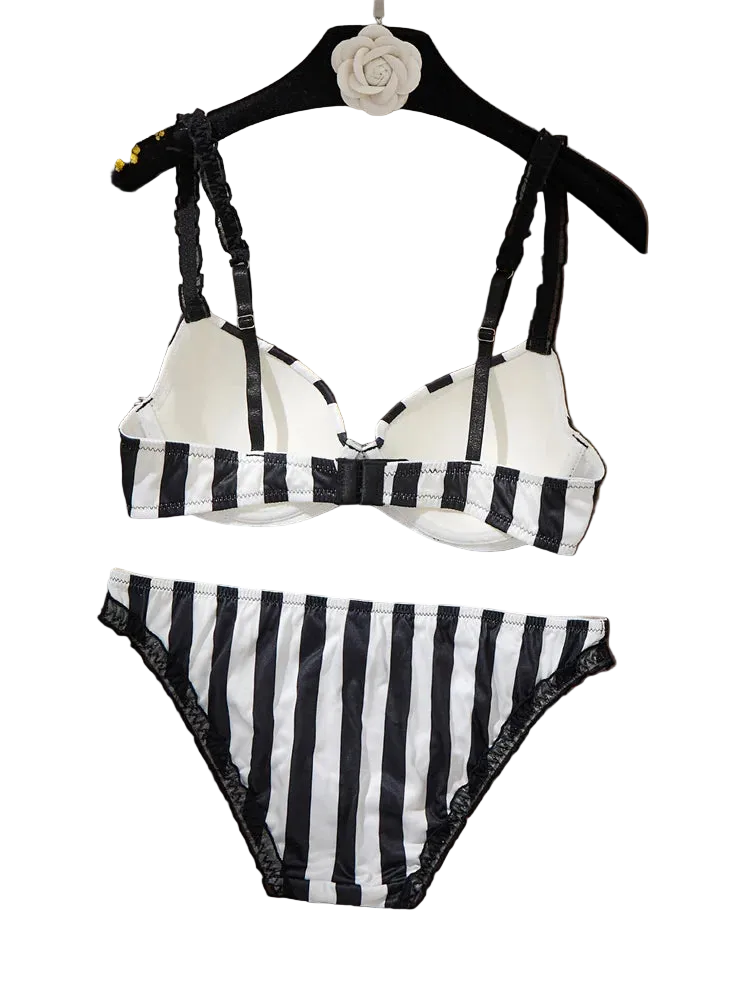 Black and white striped two-piece bikini set with lace trim, displayed on a black hanger with a white fabric backdrop and dried flowers on a wooden surface.