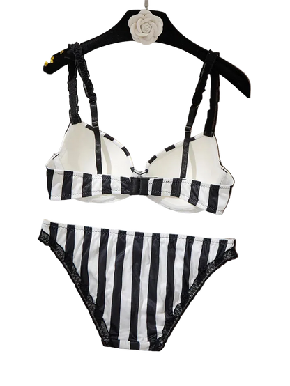 Black and white striped two-piece bikini set with lace trim, displayed on a black hanger with a white fabric backdrop and dried flowers on a wooden surface.