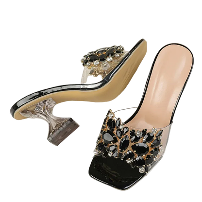 A pair of women's open-toe mule heels with clear transparent straps, glossy black square toes, and large black and silver rhinestone embellishments, displayed on a textured white fabric background.