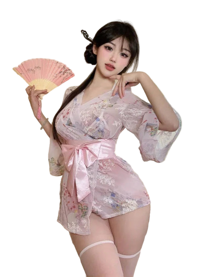 Woman in a pink and white kimono-style outfit holding a fan, with a teddy bear and pillows in the background.