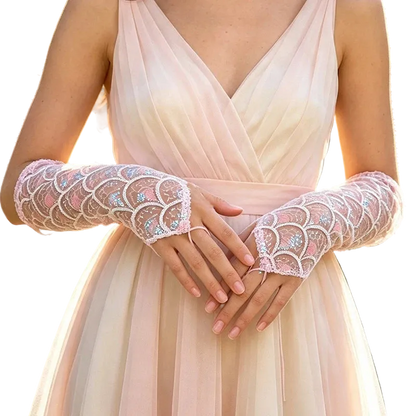 A close-up of a person wearing sheer lace opera gloves with iridescent sequin detailing over a pale blush dress, photographed outdoors with blurred greenery and pink flowers in the background.