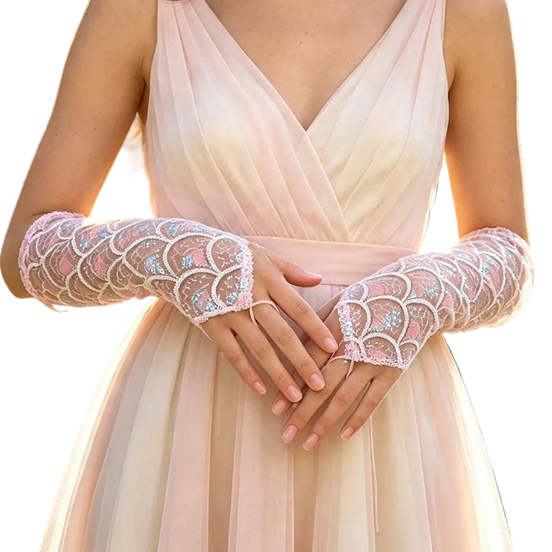 A close-up of a person wearing sheer lace opera gloves with iridescent sequin detailing over a pale blush dress, photographed outdoors with blurred greenery and pink flowers in the background.