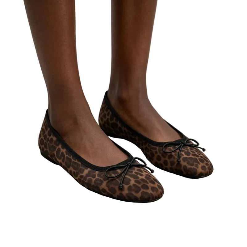 A person wearing brown and black leopard-print ballet flats with black ribbon bows, standing on light gray stone tiles.