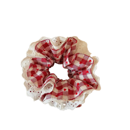 A fabric hair scrunchie with a red and white gingham pattern and a white lace trim, resting on a beige book on a wooden table.
