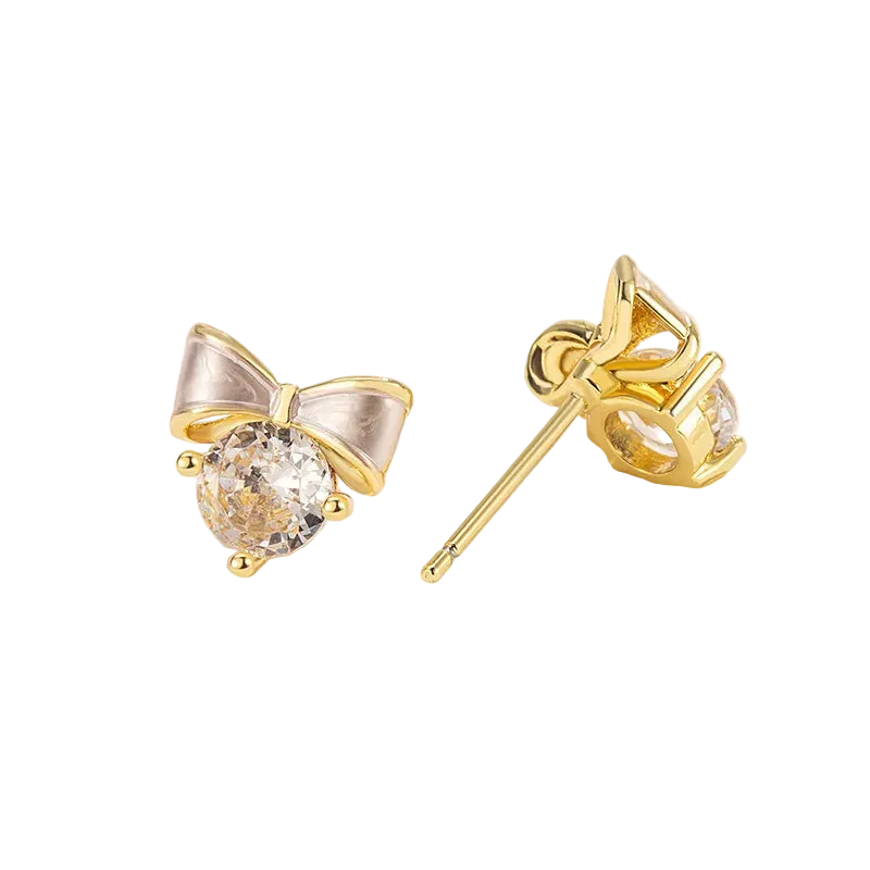 A pair of gold bow stud earrings with clear round gemstones, displayed on a white decorative surface. One earring is shown from the front, and the other is turned to show the post and back setting.