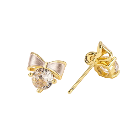 A pair of gold bow stud earrings with clear round gemstones, displayed on a white decorative surface. One earring is shown from the front, and the other is turned to show the post and back setting.