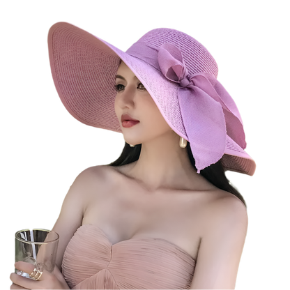 Woman wearing a pink sun hat with a bow, sitting outdoors among green plants.