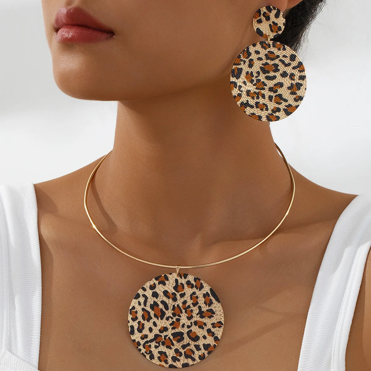 A close-up of a model wearing a gold-tone necklace and matching drop earrings with a leopard print pattern. The jewelry features a beige, brown, and black animal print on a gold-tone metal setting.