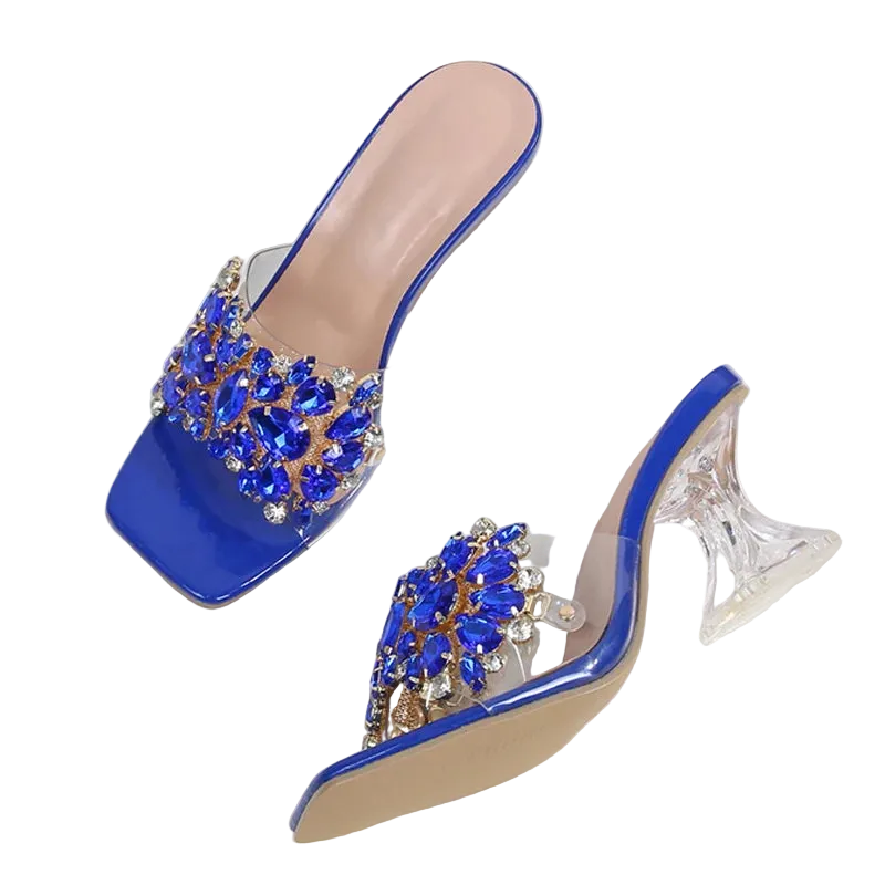 A pair of women's mule high heels with glossy blue patent leather uppers adorned with large blue and clear rhinestone embellishments, clear block heels, and beige insoles, displayed on a white fabric surface.
